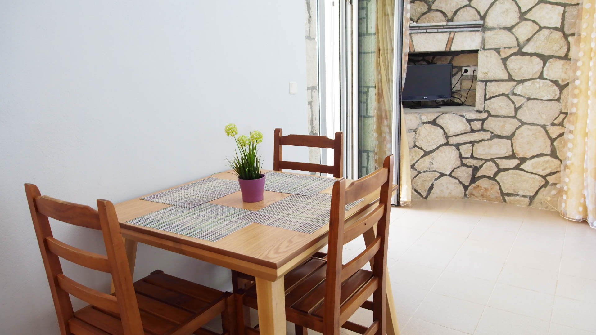 Three wooden chairs and a table, a small tv and a stone wall behind.